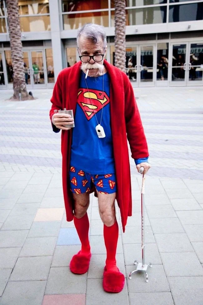 Ảnh hài hước về người đàn ông lớn tuổi mặc trang phục Superman, áo choàng đỏ, dép lông và chống khung tập đi ngoài quảng trường