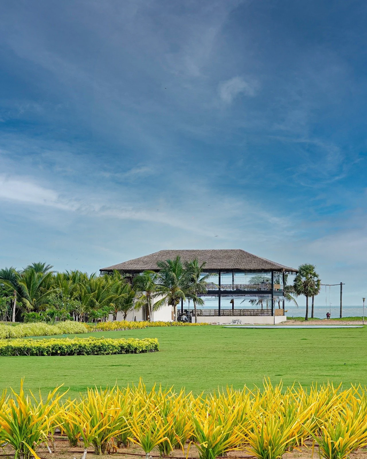 Mẫu nhà 2 tầng đẹp phong cách nghỉ dưỡng nhiệt đới nằm bên bờ biển, surrounded by cây xanh và bãi cỏ.